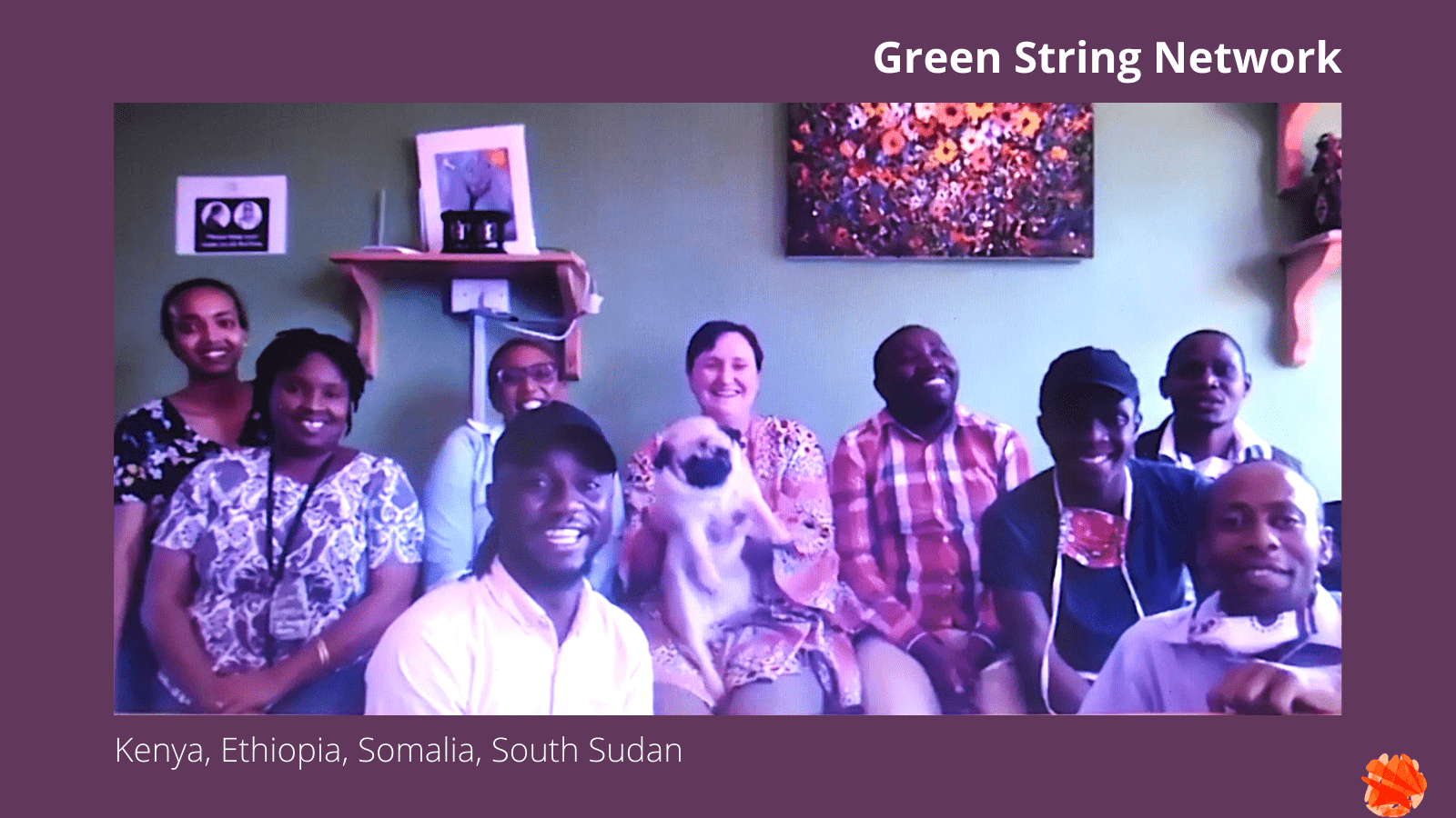 Portrait of the Green String Network team: Eigh people are sitting next to each other on a sofa, while two people crouch in front. Everyone is smiling at the camera. One woman holds a small dog in her lap. The walls behind them are a vibrant green, with paintings on them.