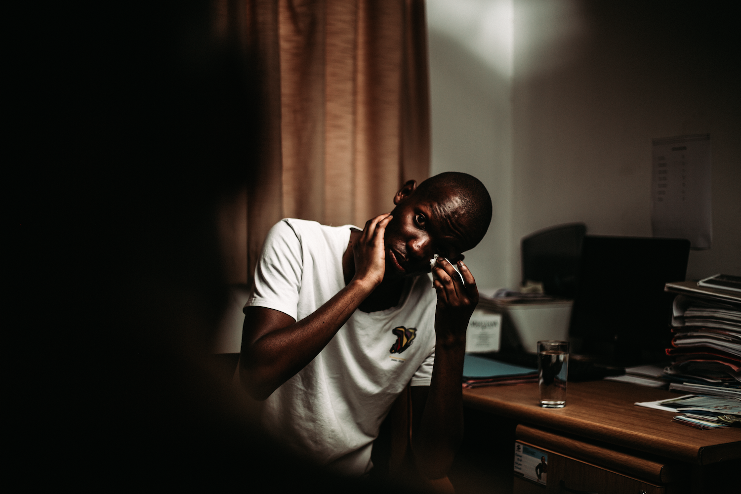 Young man sits in a therapist's room, facing a blurry figure we can't see, gazing at the camera and wiping away a tear