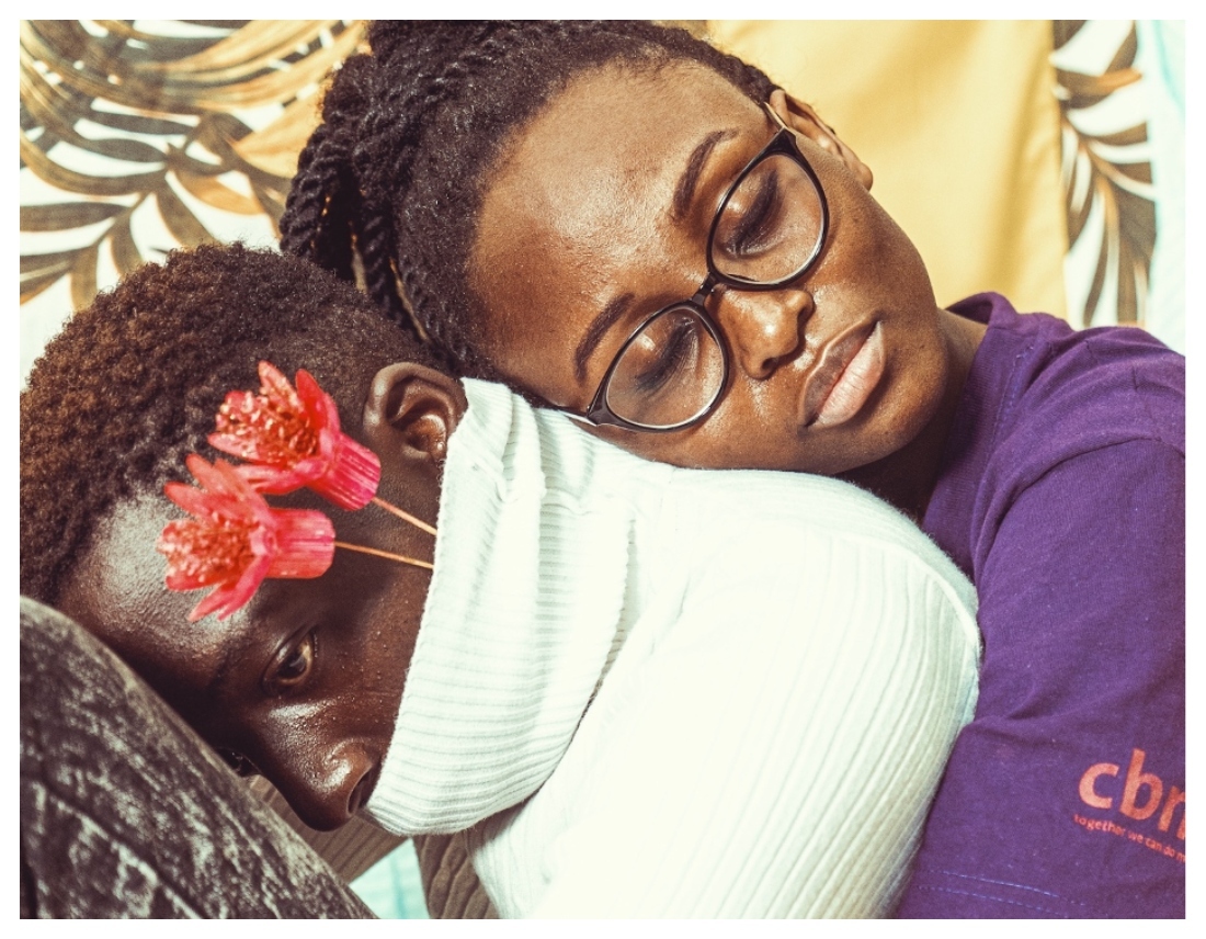 A boy has his head on his knee, with a mask on his mouth and flowers growing out of it. A young girl hugs him from behind.