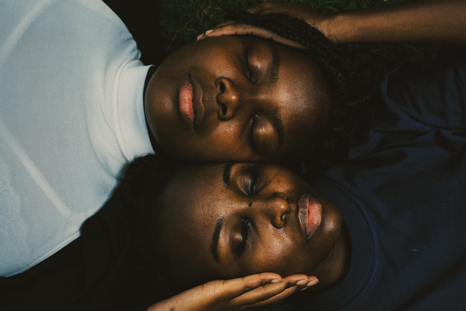 Two young women rest with their heads next to each other and eyes closed, with one hand on the other's cheek.