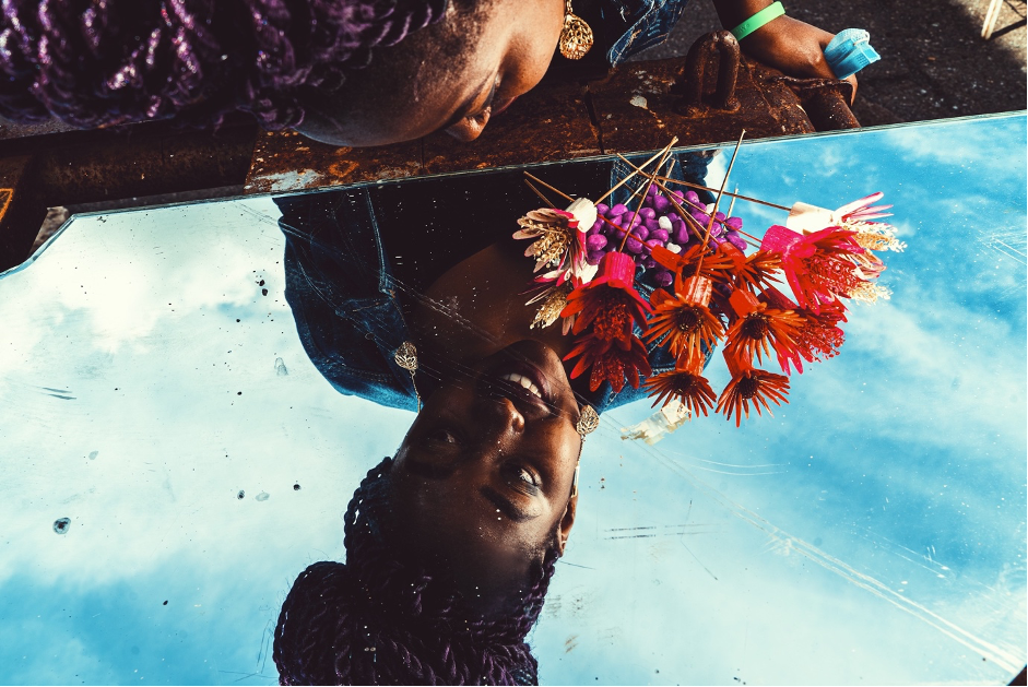 A young girl holding colourful flowers against the blue sky in background looking at her reflection in the mirror