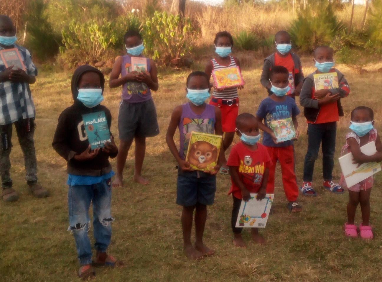 A group of young people stand in a sunny, grassy space each holding a picture book and looking at the camera