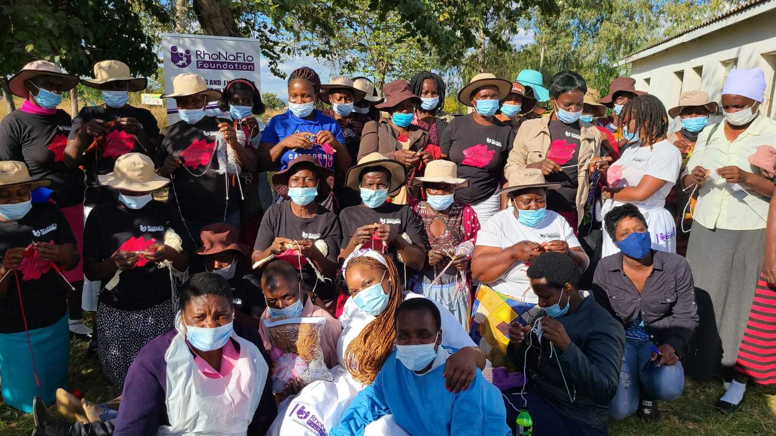 Large group of black women wearing face masks smile at the camera, and hold a banner saying 'Rhonaflo'