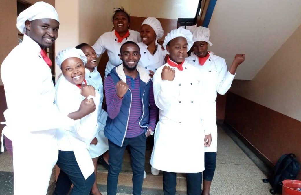 A group of young Kenyans wearing chef coats and hats smile at the camera, flexing their arms in a sign of strength