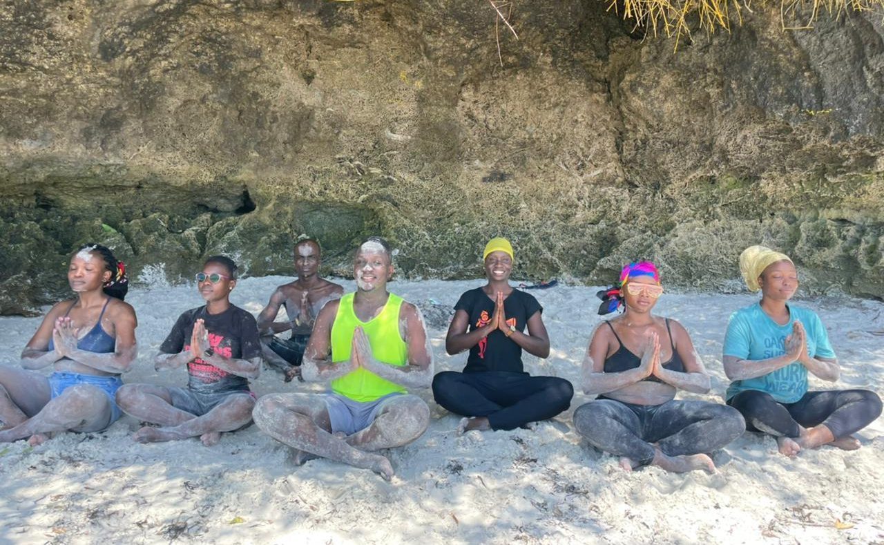 Seven women sit cross-legged, with their hands in a prayer pose, meditating